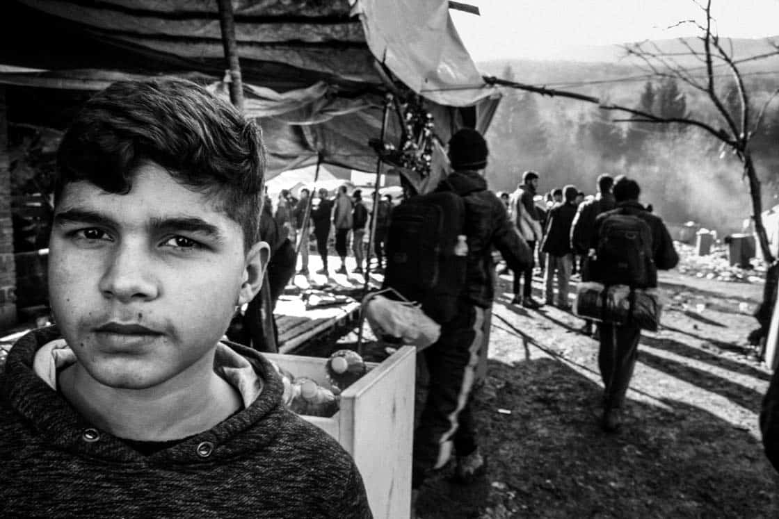 5 – Camp Vucjak, Herbst 2019. Mit 12 Jahren ist er einer der Jüngsten auf der Müllhalde. Der Junge war vor den Taliban in Afghanistan geflohen. Unterwegs hatte er sich einer Gruppe angeschlossen, um nicht mehr allein zu sein. Gemeinsam strandeten sie hier. Hinter ihm starten zwei Menschen „the game“. Die Schlange im Hintergrund wartet auf Essen.